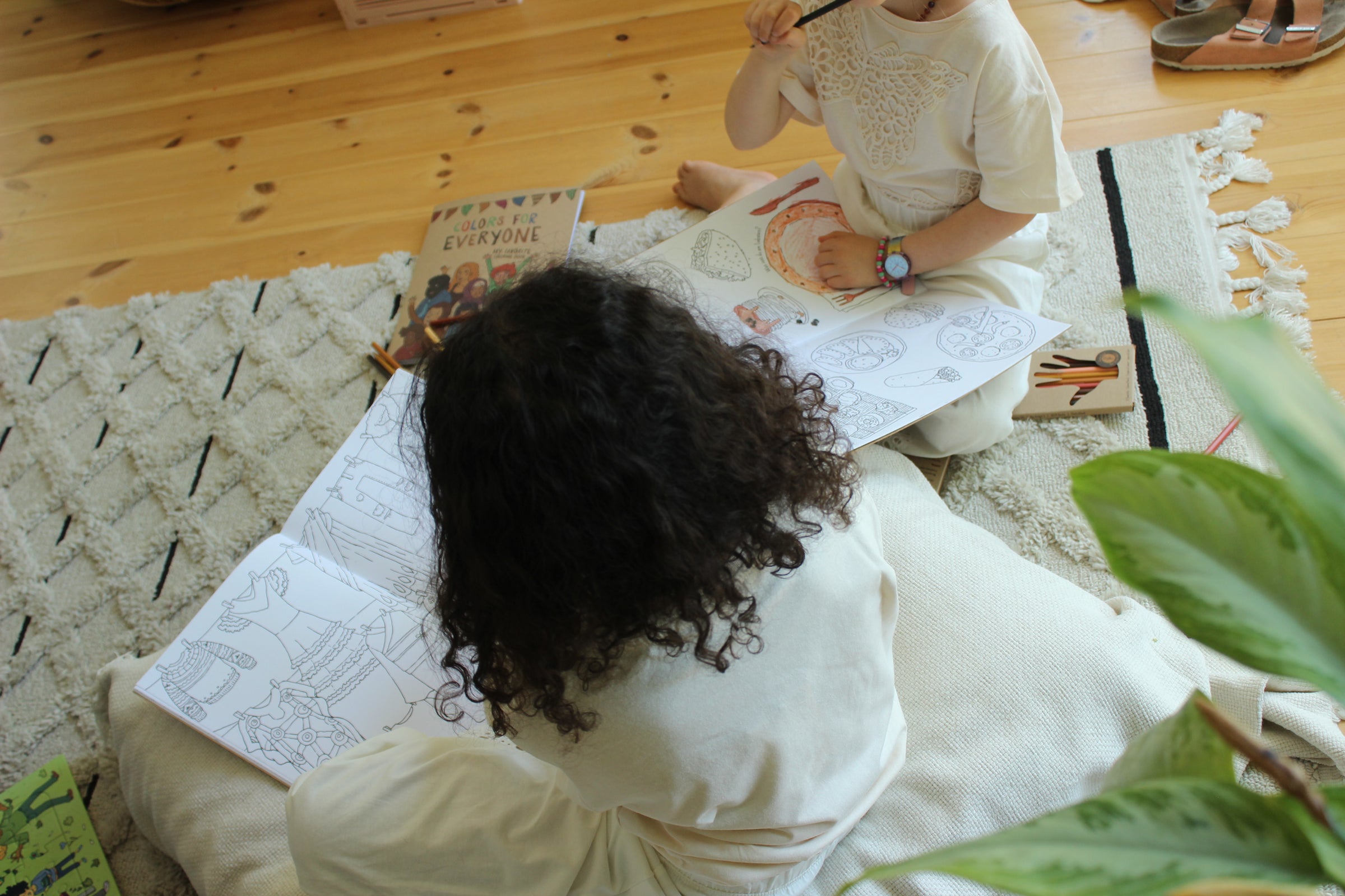 Zwei Kinder sitzen auf einem Teppich und malen gemeinsam in Malbüchern mit Hautfarben-Buntstiften
