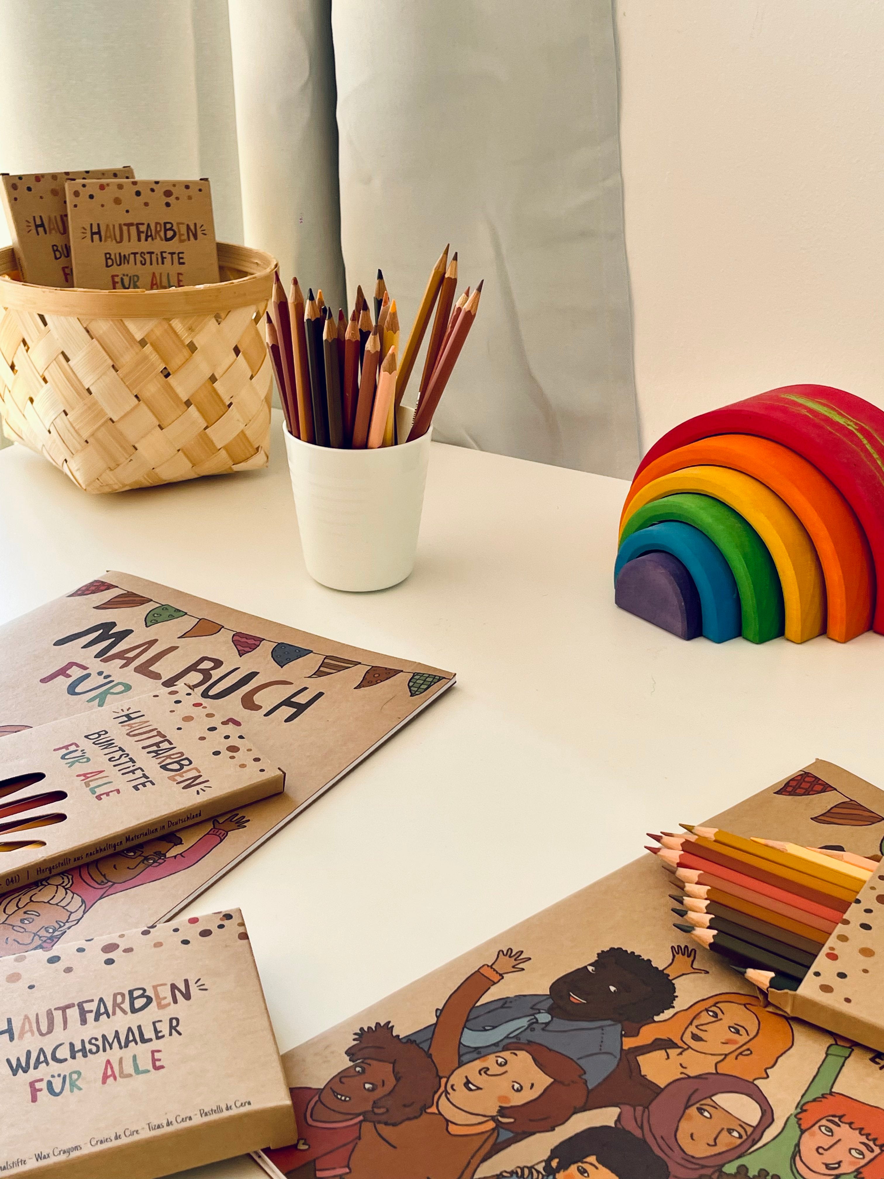 Auf dem Foto sieht man eine weiße Tischplatte. Darauf sind Malbücher, Stifte und eine Verpackung der Hautfarben-Wachsmaler zu sehen. Außerdem ein bunter Holzregenbogen.