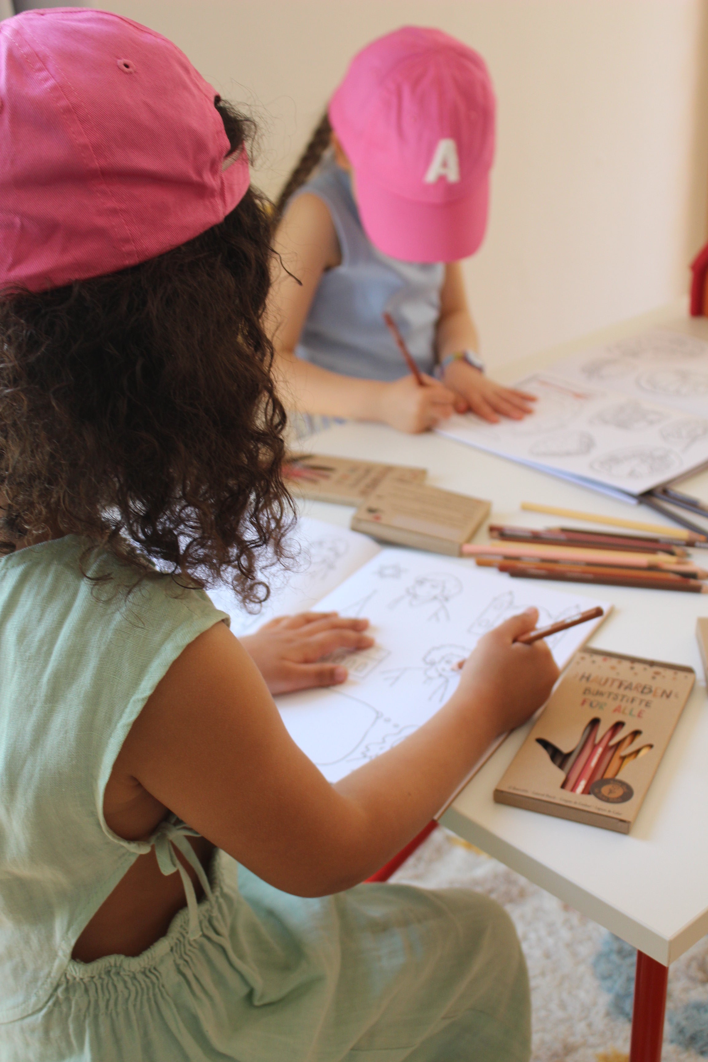 Das Foto zeigt zwei Kinder an einem Schreibtisch. Beide malen mit den Hautfarben Buntstiften in ein Hautfarben Malbuch für alle.