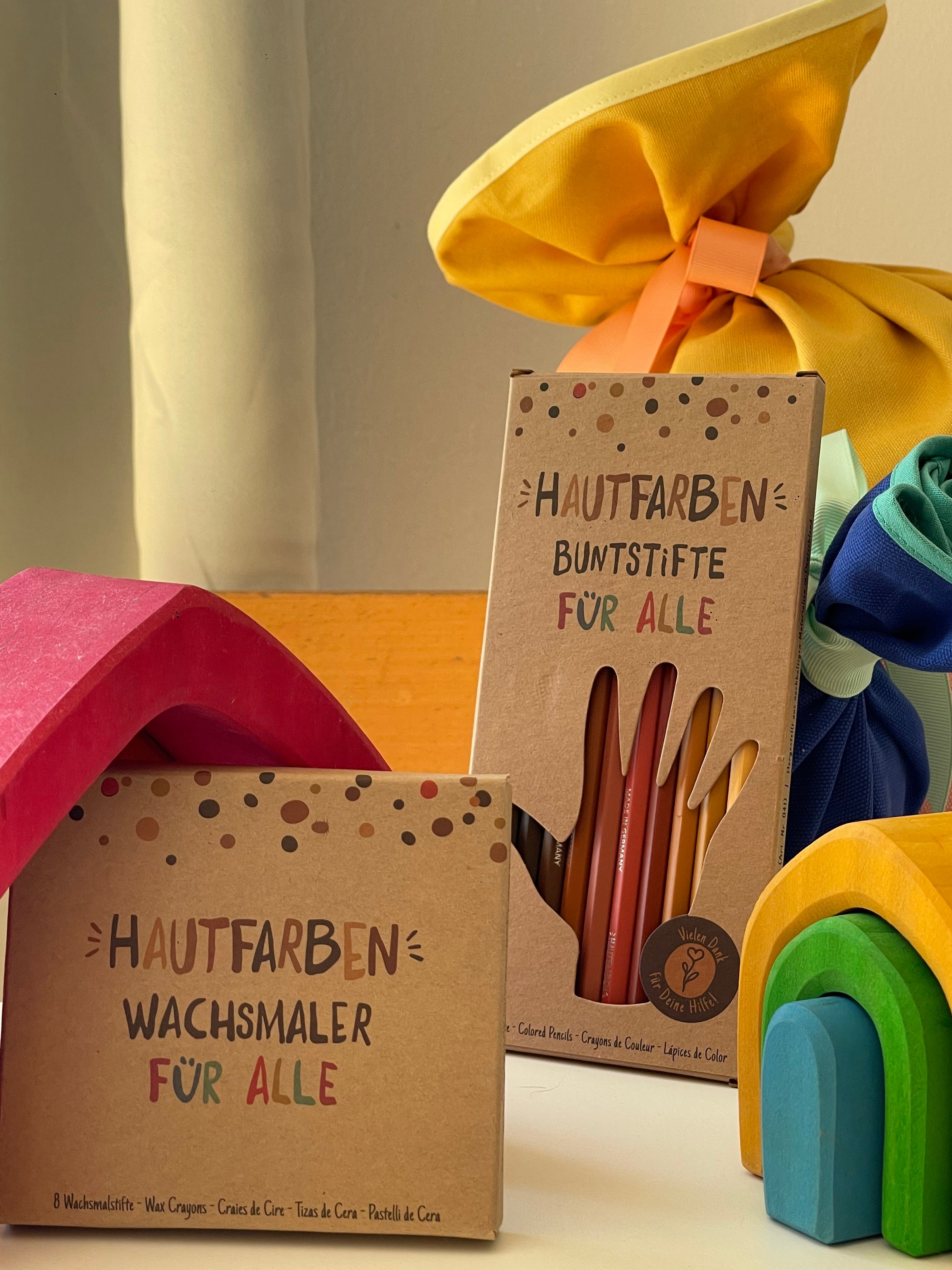 Im Vordergrund sieht man eine Verpackung Hautfarben Wachsmalstifte. Die Verpackung lehnt an einem kleinen roten Holzhaus. Dahinter steht eine Verpackung Hautfarben Buntstifte. 